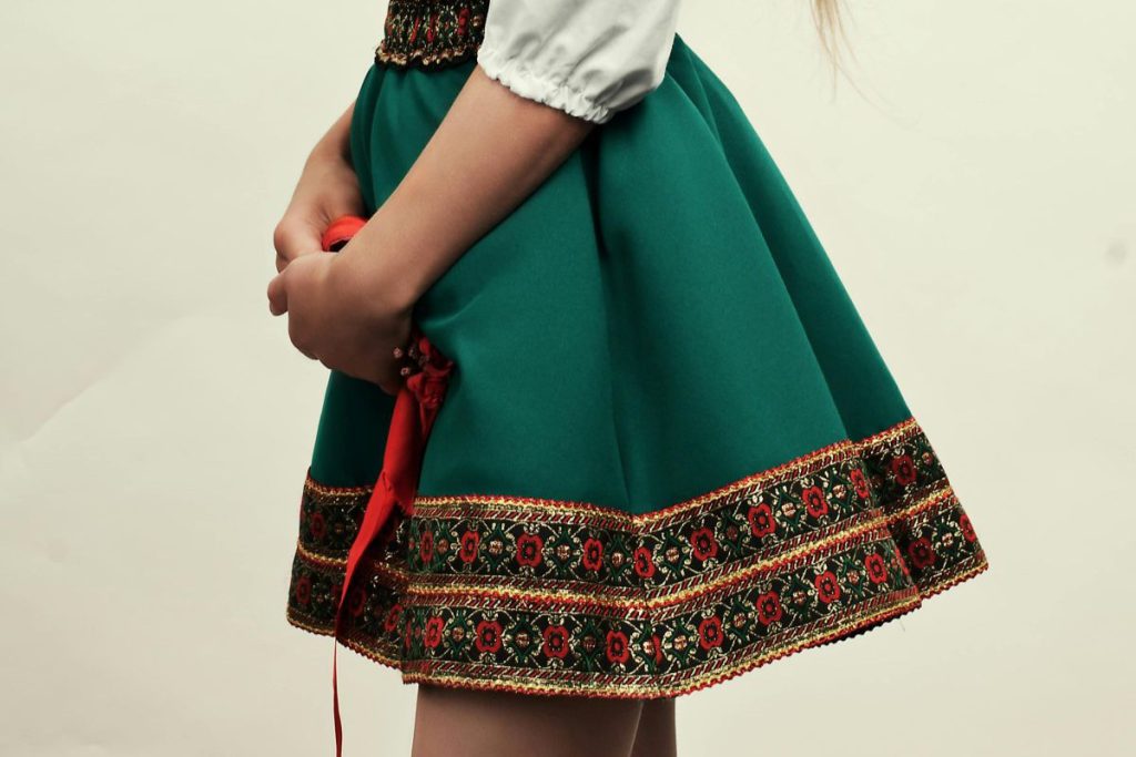 Zbliżenie na dłonie osoby trzymającej czerwoną wstążkę na tle stroju ludowego; głównym elementem jest ciemnozielona, rozkloszowana spódnica folk wykończona na dole szerokim pasem z haftowanymi czerwonymi kwiatami.